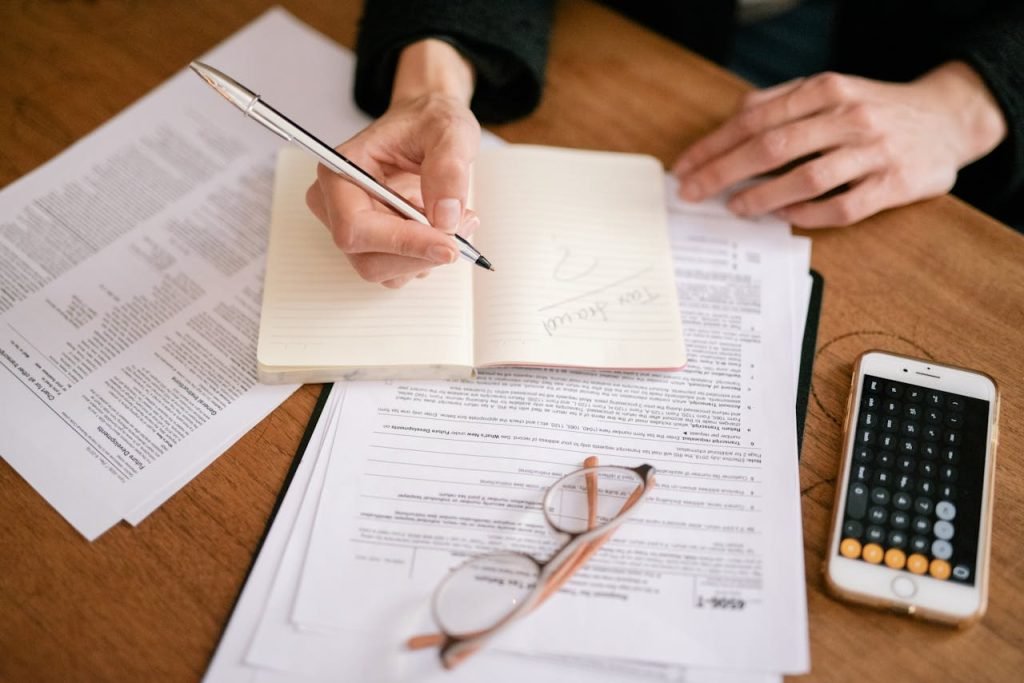 Close-up of a person writing in a notebook with documents and a calculator, managing finances.
